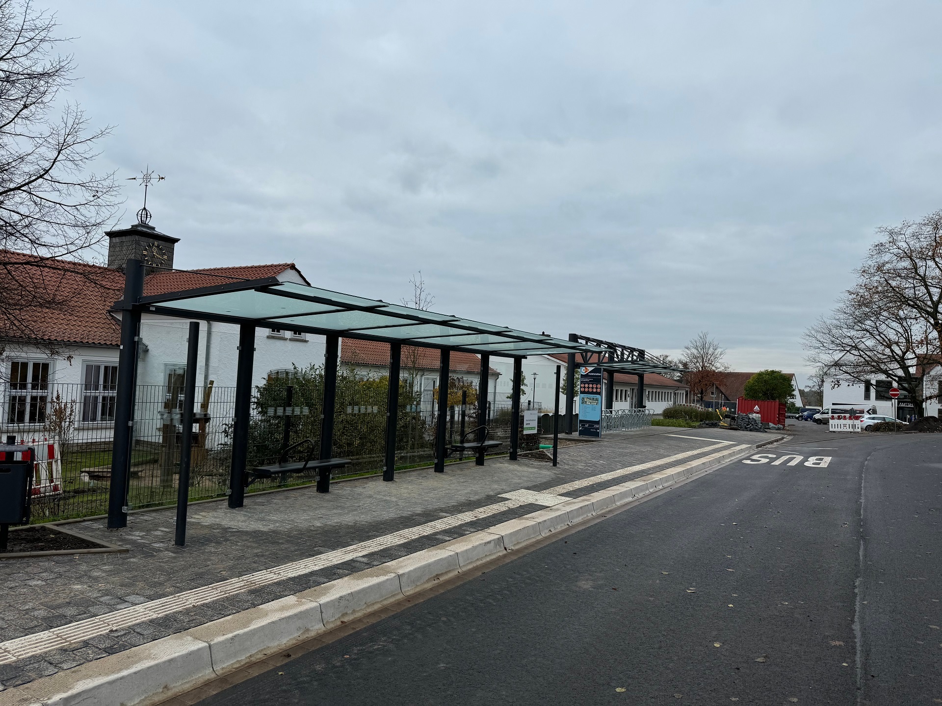 Eine Bushaltestelle mit einem erhöhten, weißen Bordstein, einem gläsernen Unterstand für Fahrgäste, vor einem weißen Gebäude.