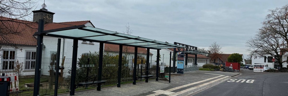 Eine Bushaltestelle mit einem erhöhten, weißen Bordstein, einem gläsernen Unterstand für Fahrgäste, vor einem weißen Gebäude.