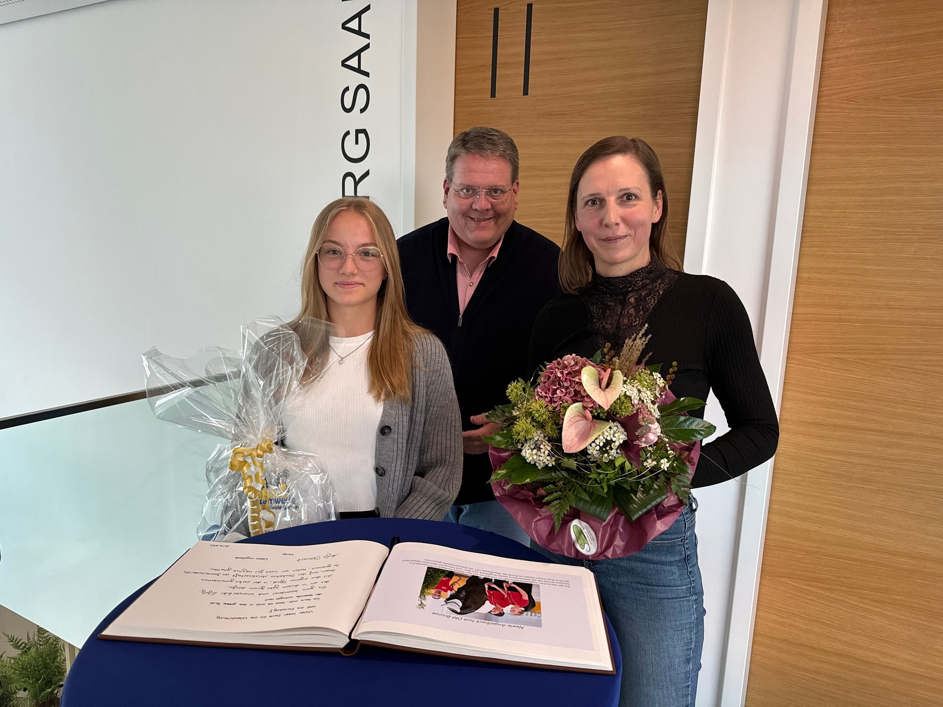 Zwei junge Frauen stehen an einem Stehtisch vor einem aufgeschlagenem Gästebuch. Im Hintergrund steht der Bürgermeister. Die beiden Frauen halten Blumen und ein Präsent in den Händen.