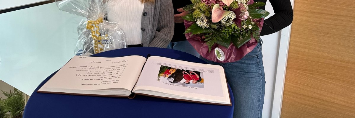 Zwei junge Frauen stehen an einem Stehtisch vor einem aufgeschlagenem Gästebuch. Im Hintergrund steht der Bürgermeister. Die beiden Frauen halten Blumen und ein Präsent in den Händen.