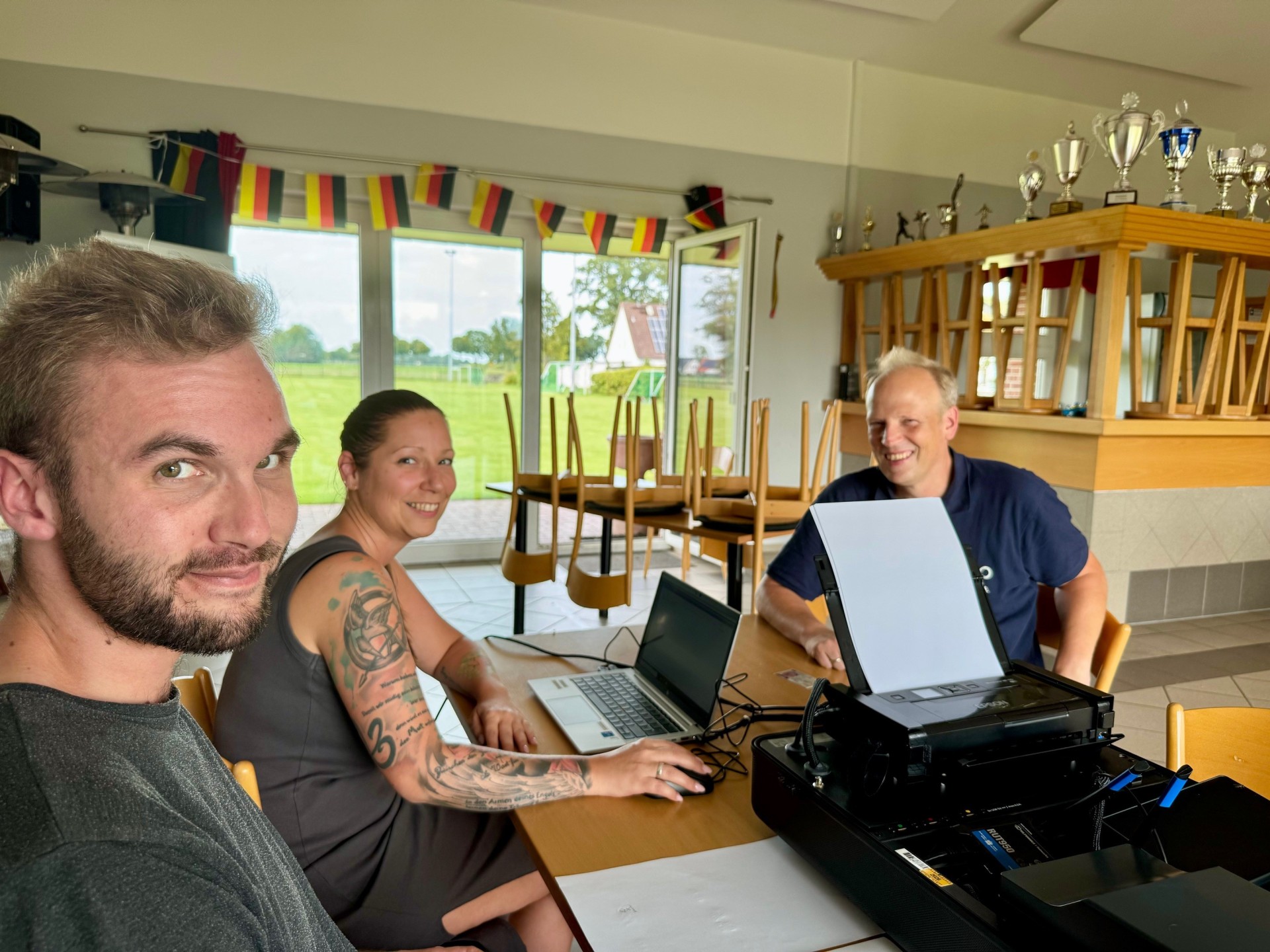 Eine Frau und ein Mann, Mitarbeiter der Gemeinde Stemwede, sitzen an einem Tisch in einem Vereinsheim und bearbeiten an einem Laptop ein Anliegen eines Bürgers, der ihnen gegenüber sitzt und für das Fot freundlich in die Kamera lacht.