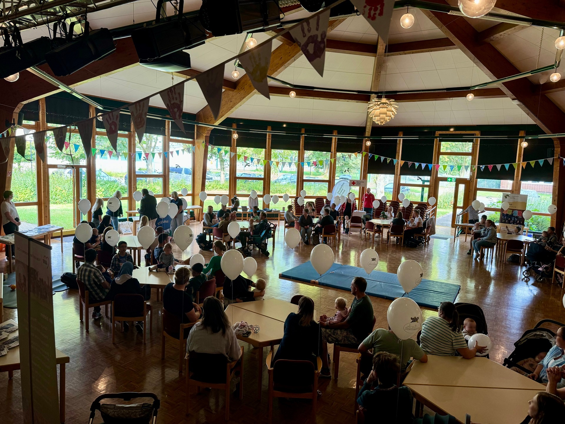Eine Veranstaltungshalle, in der zahlreiche Erwachsene mit kleinen Kindern an Tischen sitzen. Die Halle ist mit Luftballons und Girlanden geschmückt.