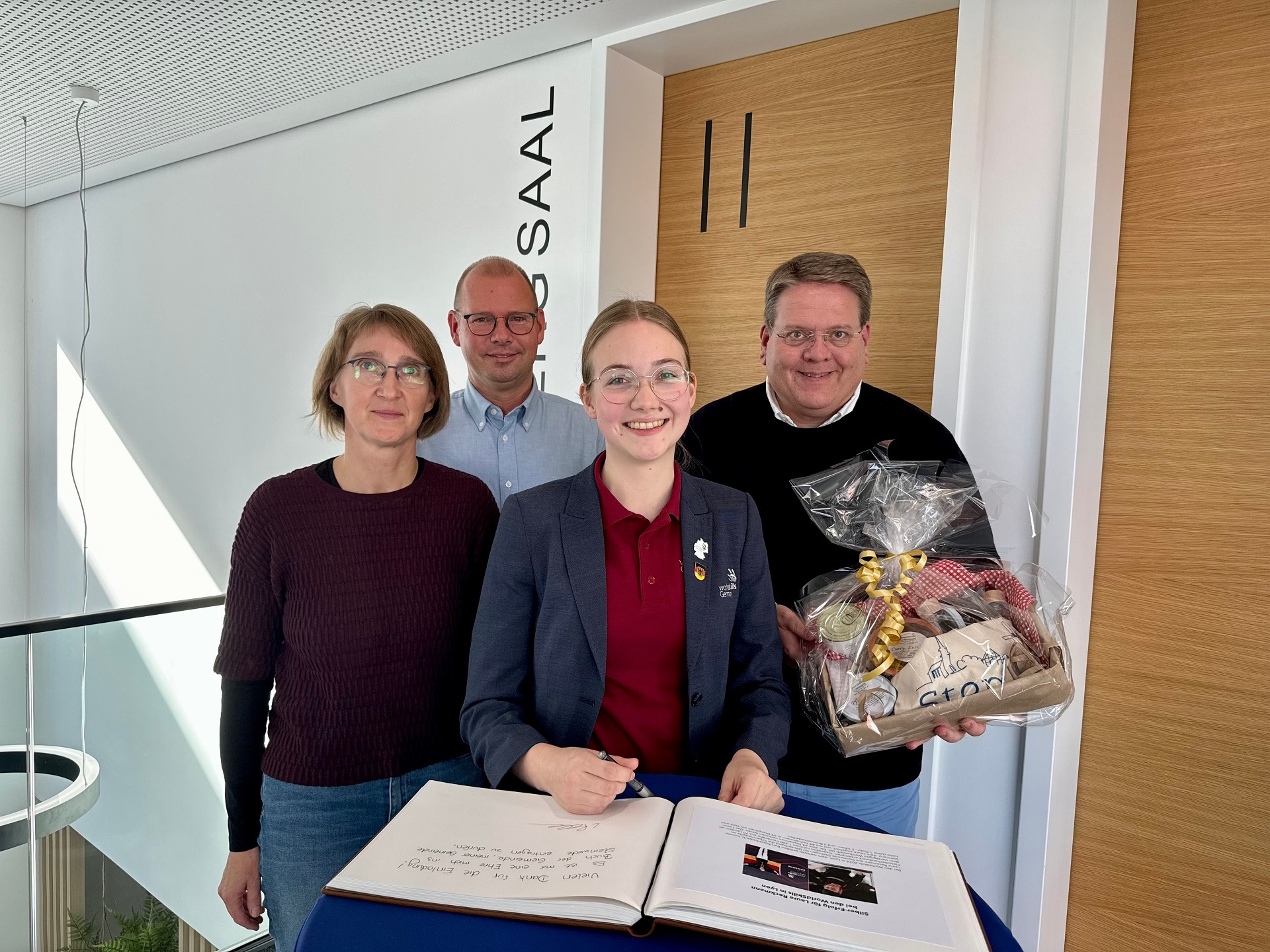 Eine junge Frau mit Brille und einem dunklen Blazer steht an einem Stehtisch und trägt sich in ein vor ihr liegendes Gästebuch ein. Hinter Ihr stehen die Eltern der jungen Frau sowie der Bürgermeister der Gemeinde Stemwede, der ein Präsent in den Händen hält.