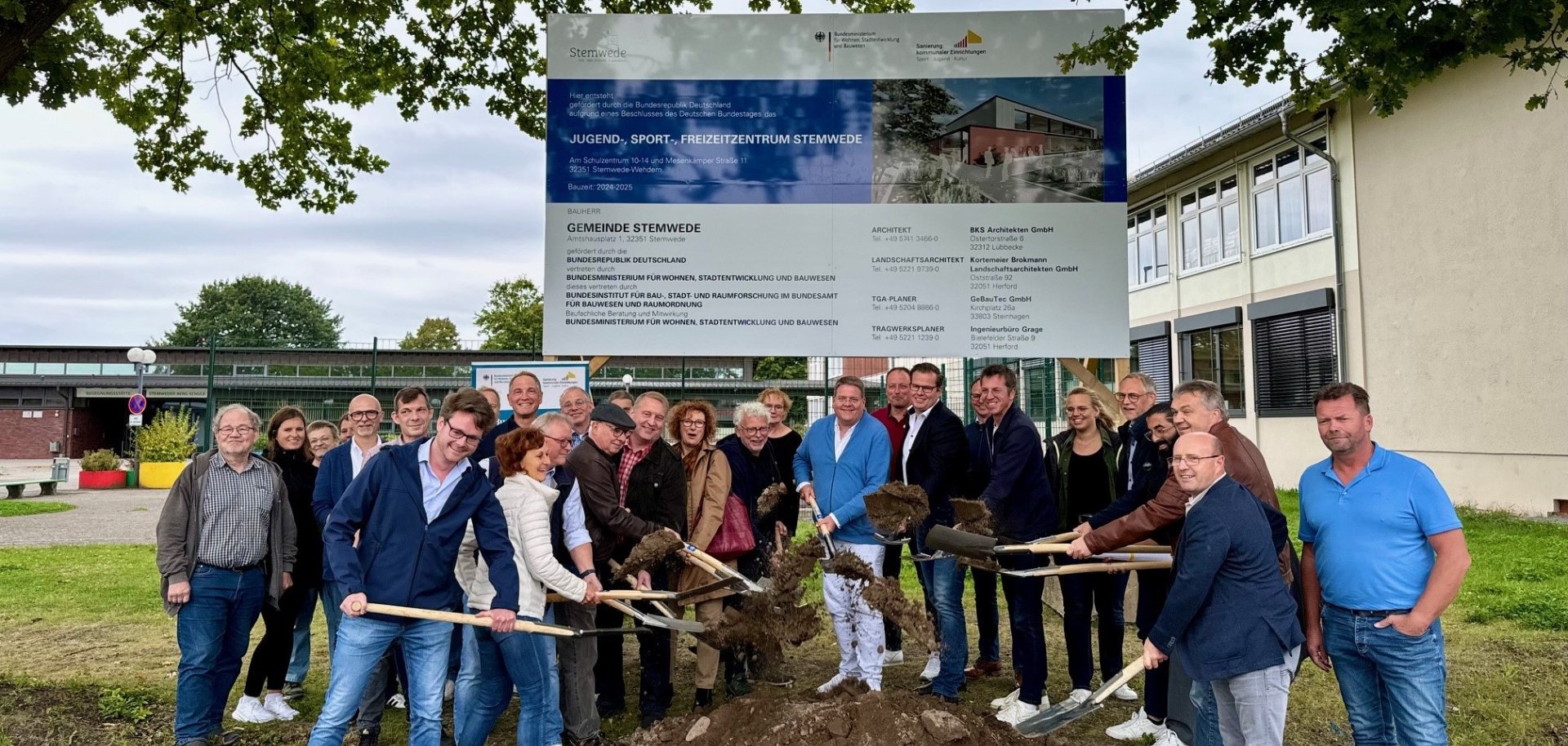 Rund 20 Personen, die unter einem großen Baustellenschild stehen. Einige Akteure haben eine Spaten in der Hand und werfen damit etwas Erde in die Höhe, als Zeichen des symbolischen Spatenstichs zum Projekt.