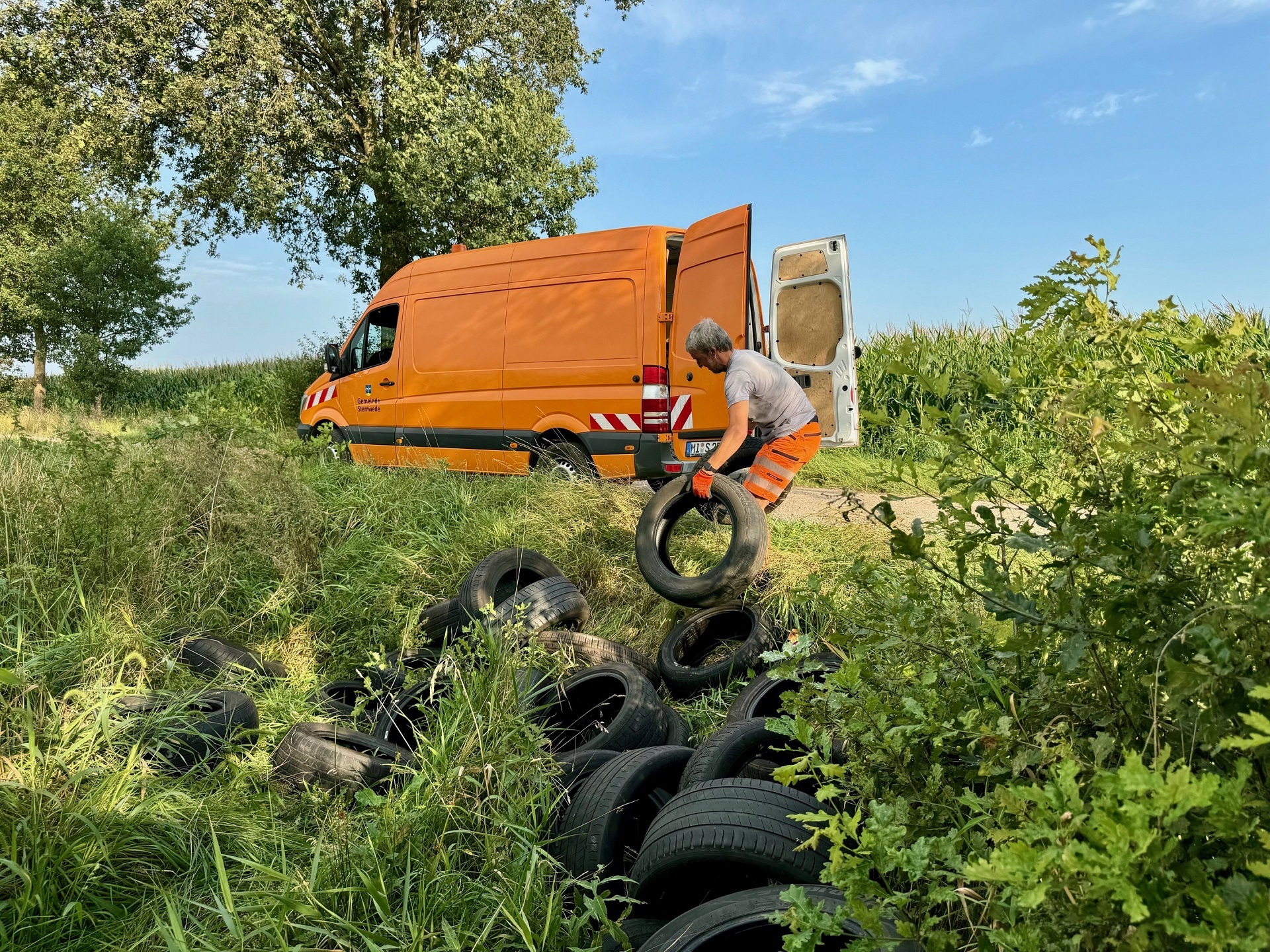 Ein Bauhofmitarbeiter der Gemeinde Stemwede ist dabei, illegal entsorgte Autoreifen aus dem Graben zu ziehen, um sie in einen orangen Bulli zu laden.