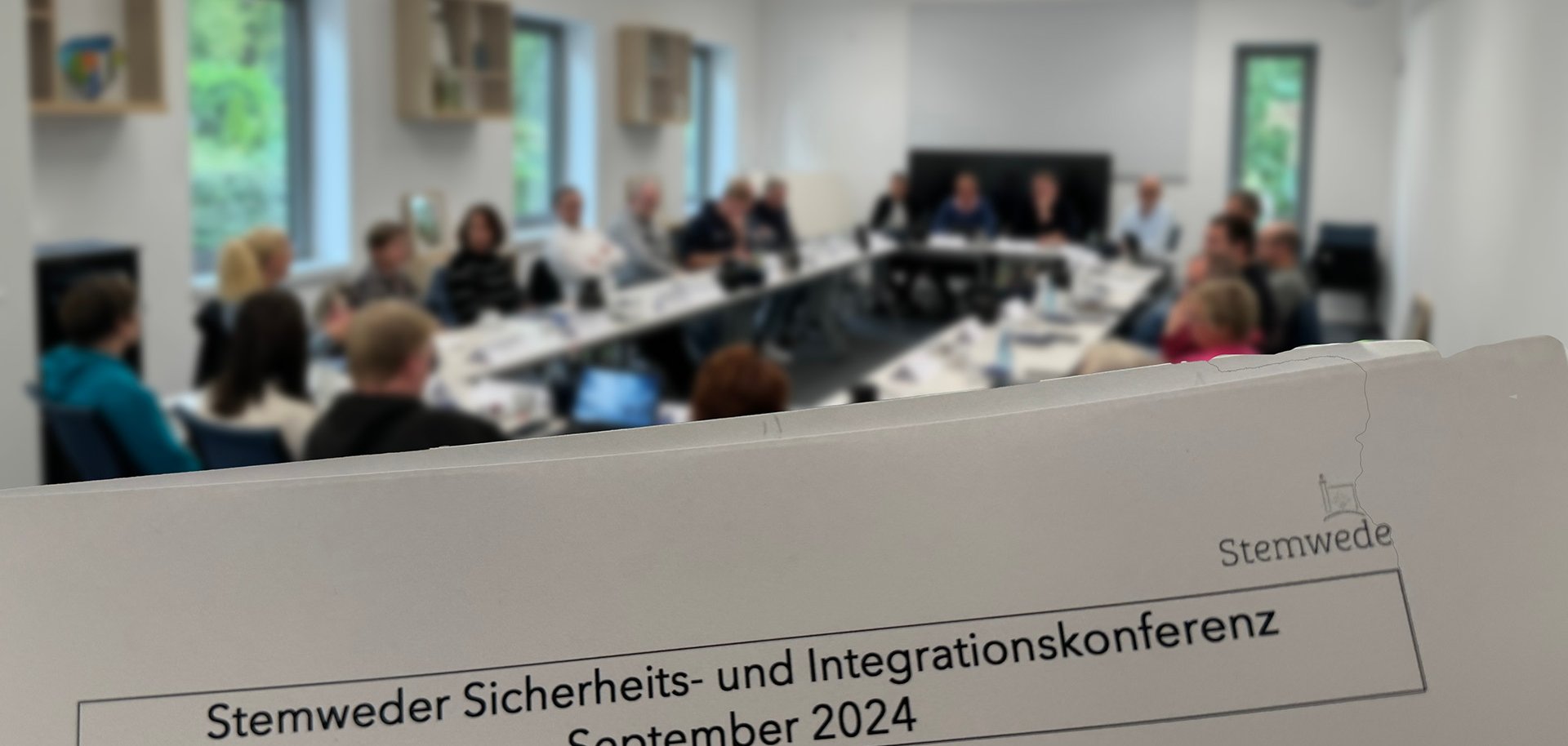 Ein nur unscharf zu erkennender Sitzungssaal in dem zahlreiche Personen sitzen. Im Vordergrund ist ein Papier zu sehen auf dem folgendes steht: Stemweder Sicherheits- und Integrationskonferenz, September 2024".