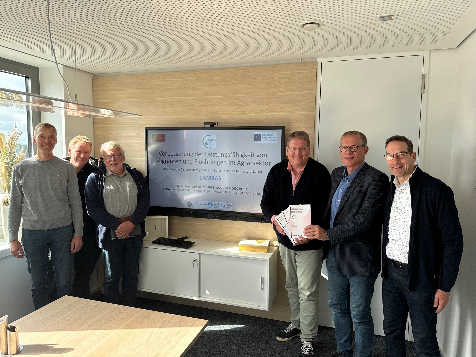 Sechs Männer, die in einem Büro vor einem Monitor stehen, auf dem ein Hinweis auf das sogenannte CAMRAS-Projekt zu lesen ist.