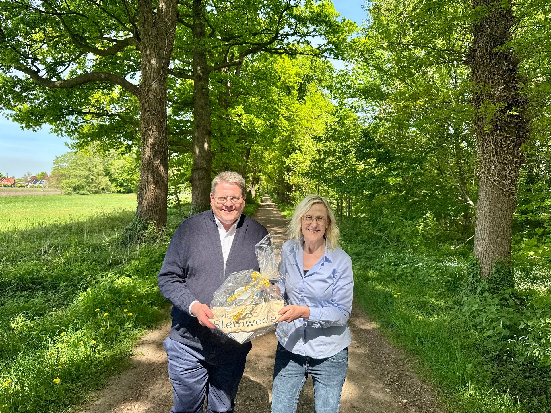 Eine Frau und ein Mann, die in einer grün blühenden Allee stehen und gemeinsam ein Präsent in die Kamera halten.