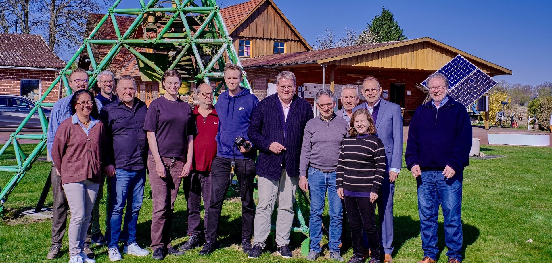 Insgesamt 13 Personen, die sich am Fuße eines technischen Gerätes mit grünem Gestänge, für ein Gruppenfoto aufgestellt haben. Im Hintergrund sind ein Gebäude sowie ein Windrad zu erkennen.