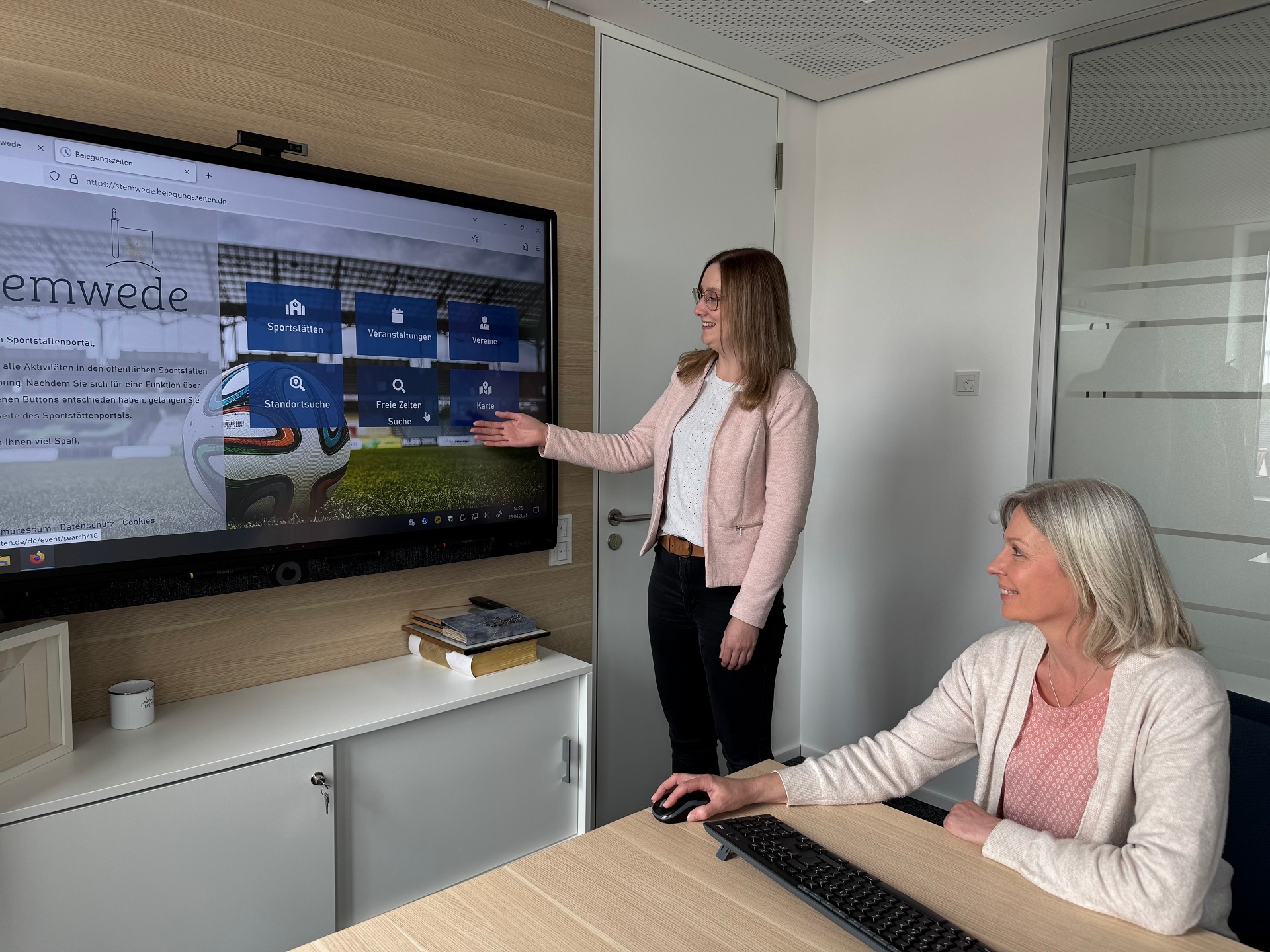 Eine Frau steht an einem großen Fernseher, der an einer Wand hängt, eine andere Frau sitzt an einem Tisch und schaut ebenfalls in Richtung Fernseher. Auf dem TV-Gerät ist ein Kalendermenü zu erkennen.
