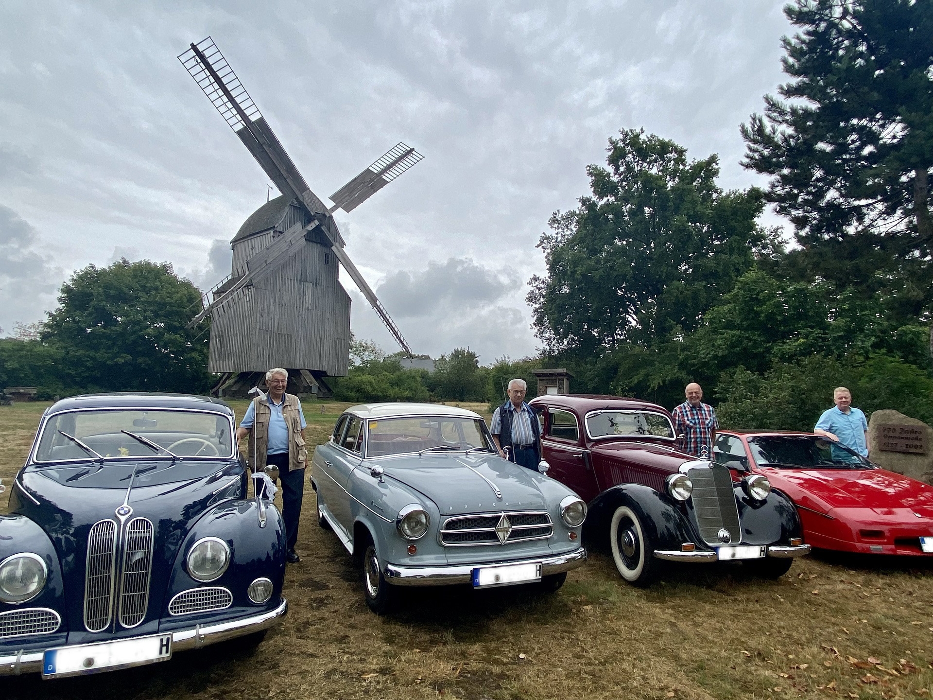 Vier Oldtimer, die samt Fahrer, vor einer hölzernen Windmühle stehen.
