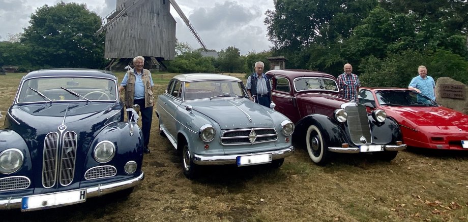 Vier Oldtimer, die samt Fahrer, vor einer hölzernen Windmühle stehen.