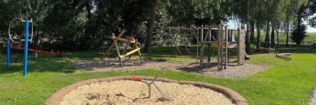 Ein Spielplatz unter Bäumen. Im Vordergrund ist ein Sandkasten zu erkennen, in dessen Mitte ein Sitzbagger aus Metall angebracht ist.