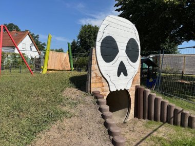 Spielplatz Oppendorf Ein Kriechtunnel auf einem Spielplatz, vor dessen Öffnung ein großer Totenkopf aus Holz hängt, so dass es den Anschein hat, dass man in den Totenkopf hineinkriechen würde.