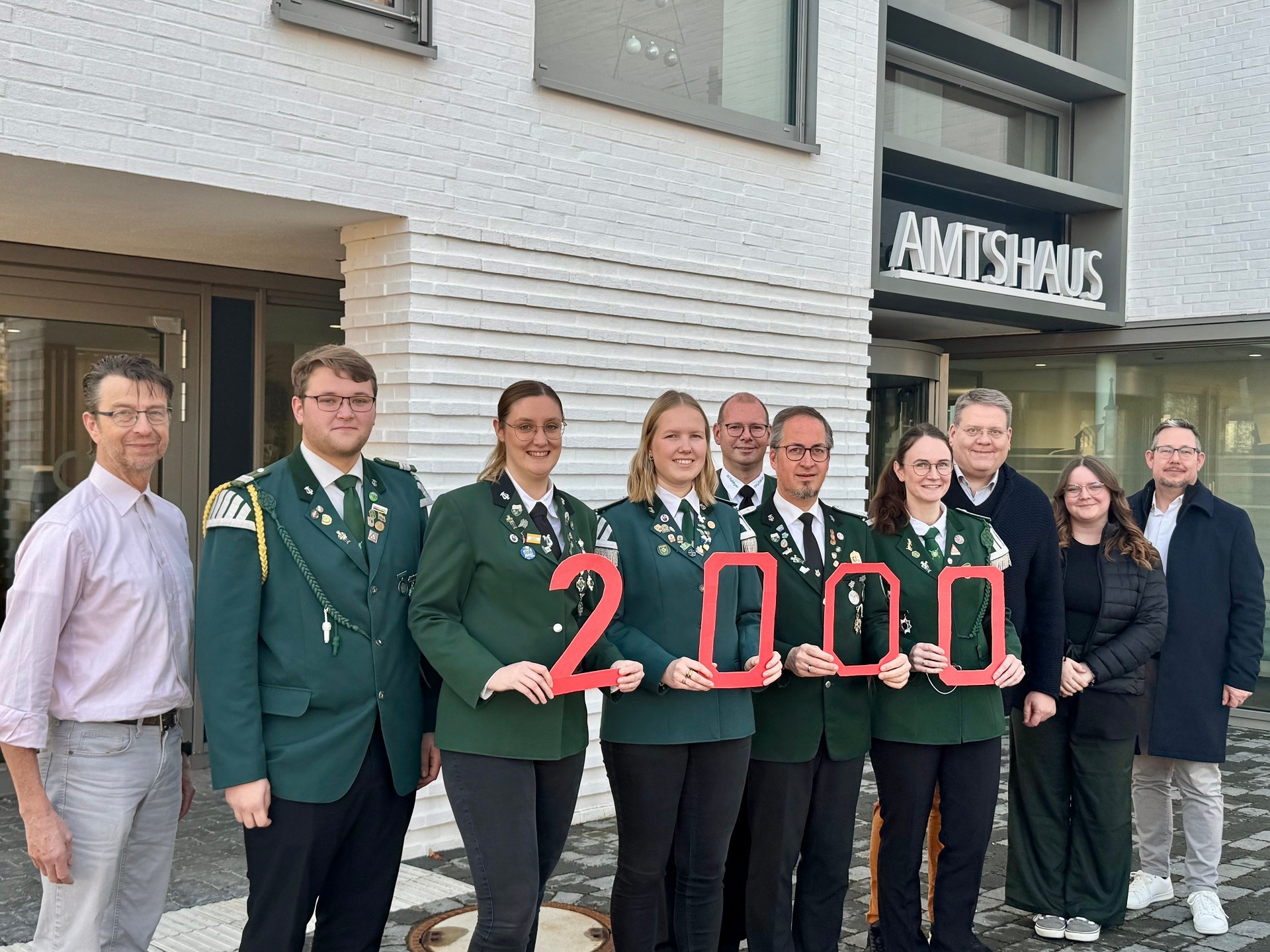 Gruppenfoto vor einem modernen Gebäude mit der Aufschrift „AMTSHAUS“. Elf Erwachsene stehen nebeneinander. In der Mitte tragen fünf Personen grüne Uniformjacken mit Abzeichen und halten rote Zahlen, die zusammen „2000“ ergeben. Die übrigen Personen tragen Alltags- oder Businesskleidung. Alle blicken in die Kamera und lächeln.