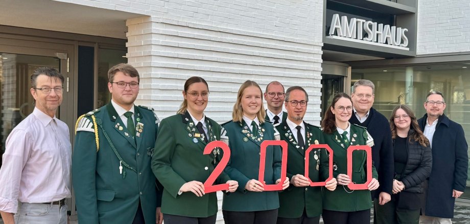 Gruppenfoto vor einem modernen Gebäude mit der Aufschrift „AMTSHAUS“. Elf Erwachsene stehen nebeneinander. In der Mitte tragen fünf Personen grüne Uniformjacken mit Abzeichen und halten rote Zahlen, die zusammen „2000“ ergeben. Die übrigen Personen tragen Alltags- oder Businesskleidung. Alle blicken in die Kamera und lächeln.