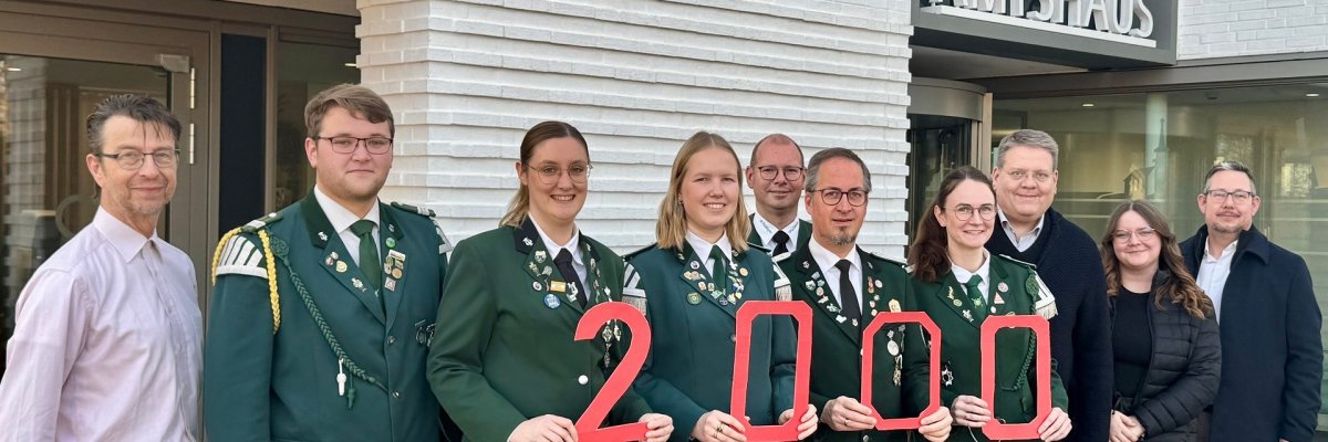 Gruppenfoto vor einem modernen Gebäude mit der Aufschrift „AMTSHAUS“. Elf Erwachsene stehen nebeneinander. In der Mitte tragen fünf Personen grüne Uniformjacken mit Abzeichen und halten rote Zahlen, die zusammen „2000“ ergeben. Die übrigen Personen tragen Alltags- oder Businesskleidung. Alle blicken in die Kamera und lächeln.