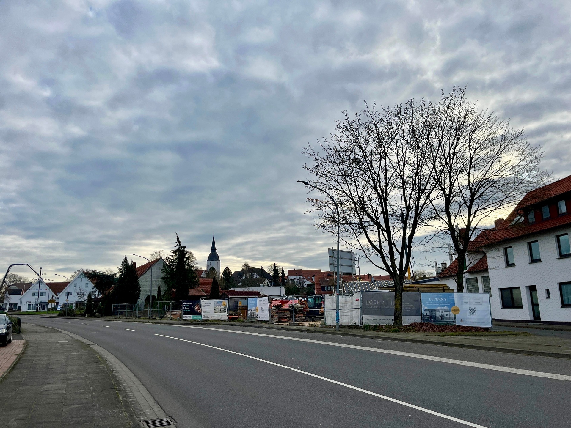 Blick auf eine ruhige Wohnstraße mit einer eingezäunten Baustelle auf der rechten Seite. Hinter Metallzäunen stehen Baucontainer, Paletten mit Baumaterial und ein gelber Minibagger. Im Hintergrund sind mehrere zweigeschossige Wohnhäuser mit Satteldächern zu sehen, Bäume ohne Laub und ein überwiegend bewölkter Himmel.
