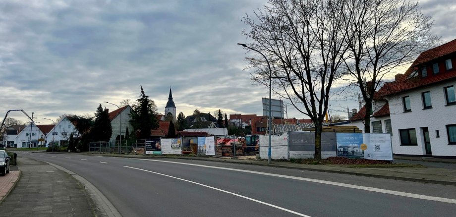 Blick auf eine ruhige Wohnstraße mit einer eingezäunten Baustelle auf der rechten Seite. Hinter Metallzäunen stehen Baucontainer, Paletten mit Baumaterial und ein gelber Minibagger. Im Hintergrund sind mehrere zweigeschossige Wohnhäuser mit Satteldächern zu sehen, Bäume ohne Laub und ein überwiegend bewölkter Himmel.
