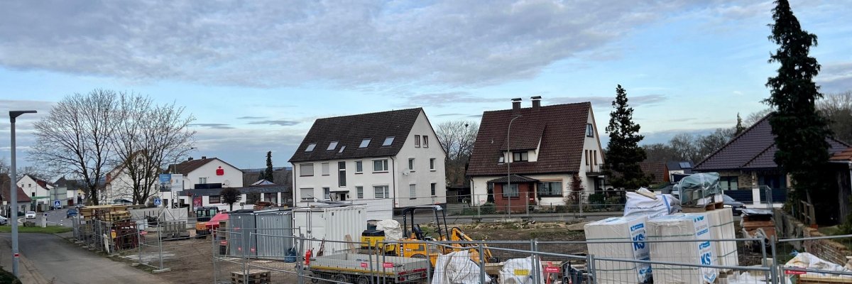 Blick auf eine ruhige Wohnstraße mit einer eingezäunten Baustelle auf der rechten Seite. Hinter Metallzäunen stehen Baucontainer, Paletten mit Baumaterial und ein gelber Minibagger. Im Hintergrund sind mehrere zweigeschossige Wohnhäuser mit Satteldächern zu sehen, Bäume ohne Laub und ein überwiegend bewölkter Himmel.