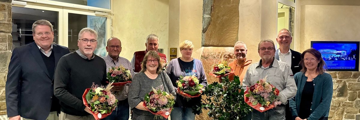 Eine Gruppe von zehn Männern und Frauen stehen in einem Eingangsbereich einer Gastronomie. Die Personen halten Blumensträuße in der Hand und haben sich für ein Gruppenfoto aufgestellt.