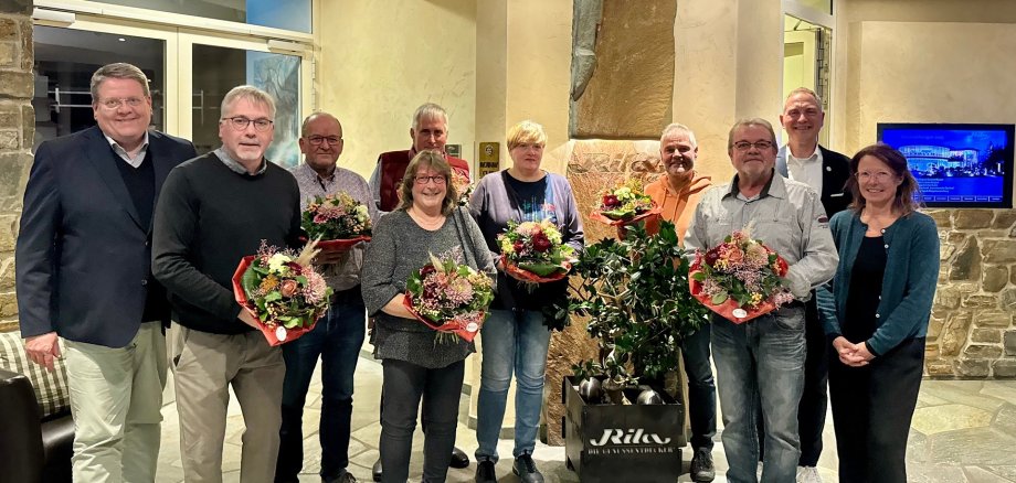 Eine Gruppe von zehn Männern und Frauen stehen in einem Eingangsbereich einer Gastronomie. Die Personen halten Blumensträuße in der Hand und haben sich für ein Gruppenfoto aufgestellt.