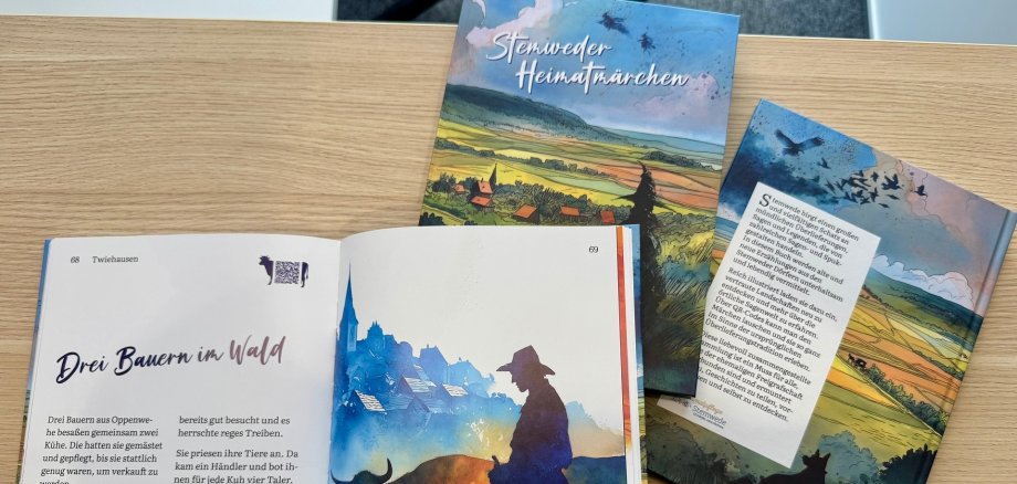 Auf einem Holztisch liegen zwei Exemplare eines bunt illustrierten Buchs mit dem Titel „Stemweder Heimatmärchen“. Ein aufgeschlagenes Buch zeigt links Text und rechts eine farbige Illustration eines Bauern mit Kuh vor einem Dorf.