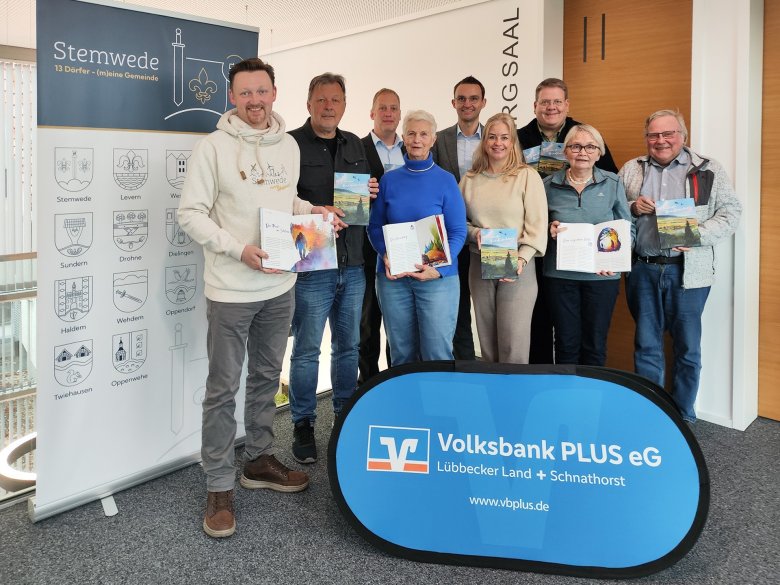 Eine Gruppe von zehn Erwachsenen steht in einem Innenraum in zwei Reihen zusammen und hält Bücher in den Händen. Vor ihnen steht ein blaues, faltbares Werbeschild der „Volksbank PLUS eG“. Links im Bild befindet sich ein großes Roll-up mit dem Schriftzug „Stemwede – 13 Dörfer (m)eine Gemeinde“ und mehreren Ortswappen. Die Personen lächeln in die Kamera und präsentieren die geöffneten oder geschlossenen Bücher. Der Raum wirkt modern, mit hellen Wänden und Holzpaneelen.