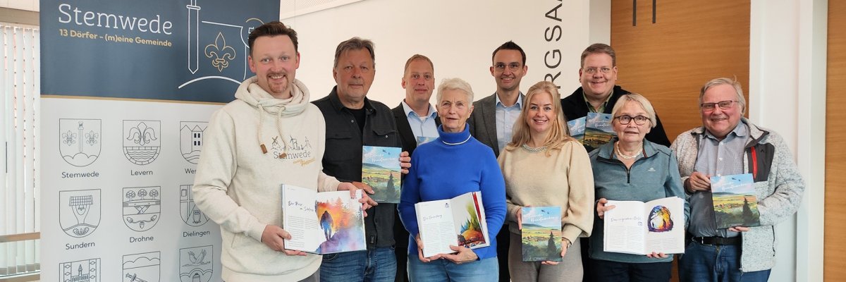 Eine Gruppe von zehn Erwachsenen steht in einem Innenraum in zwei Reihen zusammen und hält Bücher in den Händen. Vor ihnen steht ein blaues, faltbares Werbeschild der „Volksbank PLUS eG“. Links im Bild befindet sich ein großes Roll-up mit dem Schriftzug „Stemwede – 13 Dörfer (m)eine Gemeinde“ und mehreren Ortswappen. Die Personen lächeln in die Kamera und präsentieren die geöffneten oder geschlossenen Bücher. Der Raum wirkt modern, mit hellen Wänden und Holzpaneelen.
