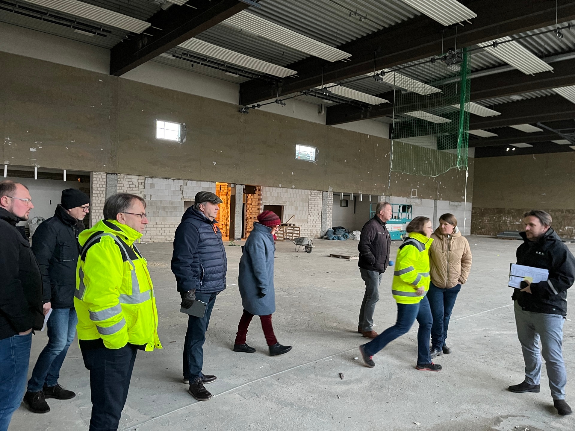 Eine Gruppe von Personen, die Winterkleidung und zum Teil auch neongelbe Jacken tragen, laufen durch eine im Bau befindliche Turnhalle.