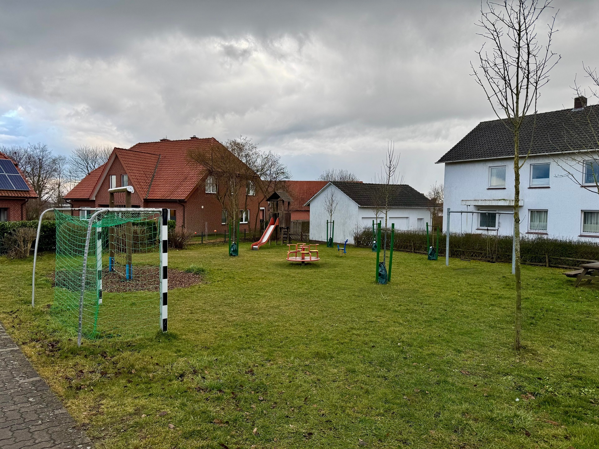 Blick auf einen Spielplatz in einer Wohnsiedlung. Es sind unter anderem ein Fußballtor, eine Schaukel, eine Rutsche und mehrere Bäume zu erkennen.
