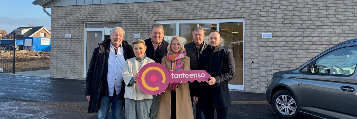 Eine Gruppe von Personen, zwei Frauen und vier Männer, die sich vor einem im Bau befindlichen Supermarktgebäude für ein Gruppenfoto aufgestellt haben. Die beiden vorne stehenden Frauen halten einen roten, überdimensionalen Schlüssel in der Hand auf dem "Tante Enso" steht.