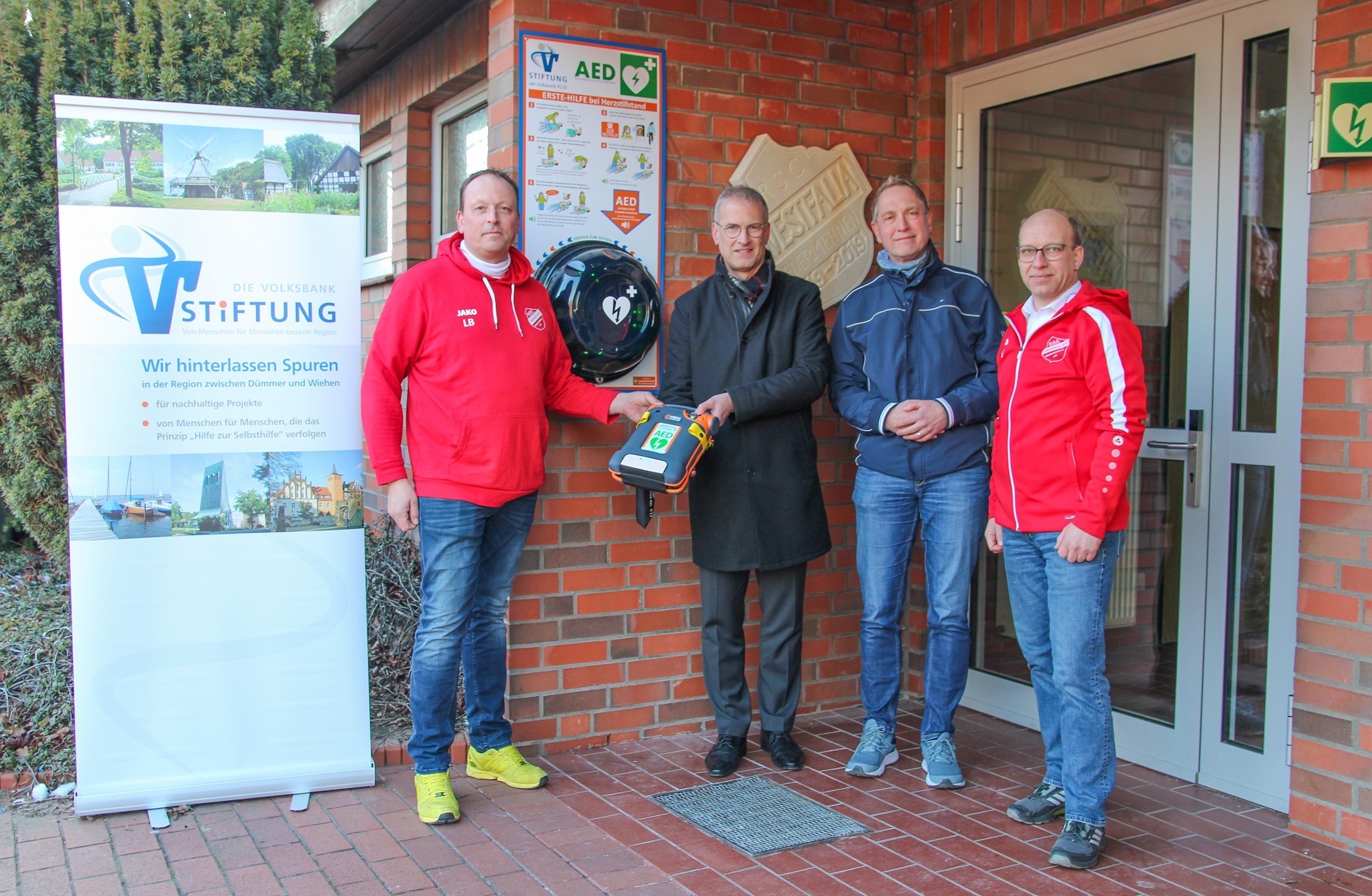 Ein Mann in einem Anzug übergibt vor dem Eingang einer Turnhalle einen Defibrillator an zwei Vereinsmitglieder des RSC Niedermehnen - zu erkennen an den roten Vereinsjacken. Ein weitere Mann in einer blauen Jacke ist ebenfalls mit dabei.