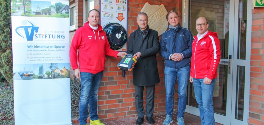 Ein Mann in einem Anzug übergibt vor dem Eingang einer Turnhalle einen Defibrillator an zwei Vereinsmitglieder des RSC Niedermehnen - zu erkennen an den roten Vereinsjacken. Ein weitere Mann in einer blauen Jacke ist ebenfalls mit dabei.