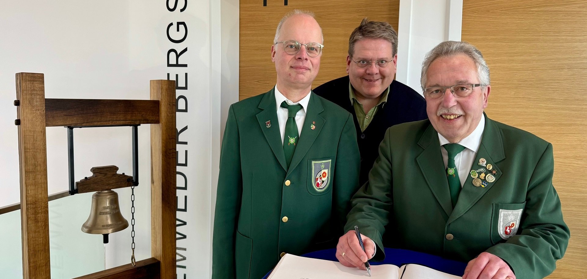 Zwei Männer in grüner Schützenuniform und der Bürgermeister der Gemeinde Stemwede stehen an einem blauen Stehtisch. Einer der beiden Schützen trägt sich in ein Gästebuch ein.