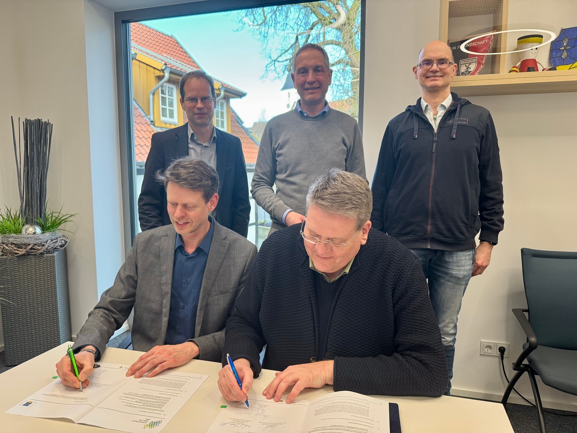 Zwei Männer sitzen nebeneinander an einem Tisch und unterzeichnen einen Vertrag. Hinter den beiden stehen drei weitere Männer und lächeln in die Kamera.