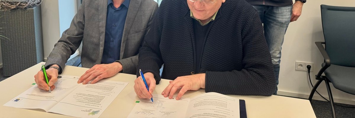 Zwei Männer sitzen nebeneinander an einem Tisch und unterzeichnen einen Vertrag. Hinter den beiden stehen drei weitere Männer und lächeln in die Kamera.