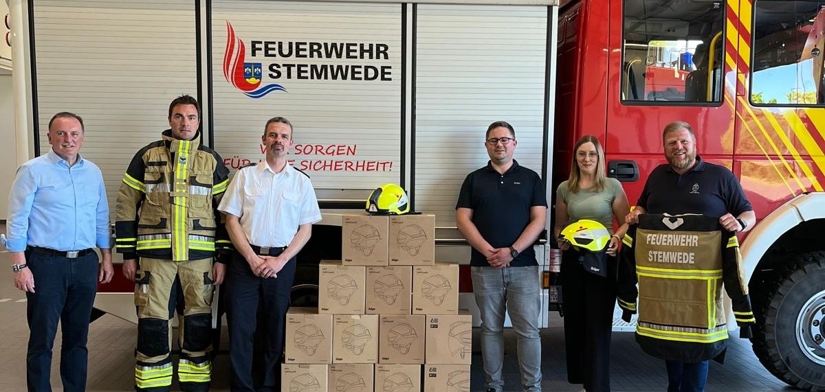 Mehrere Personen, die vor einem Feuerwehrauto der Feuerwehr Stemwede stehen und neongelbe Helme sowie sandfarbende Uniformen in den Händen halten und präsentieren.
