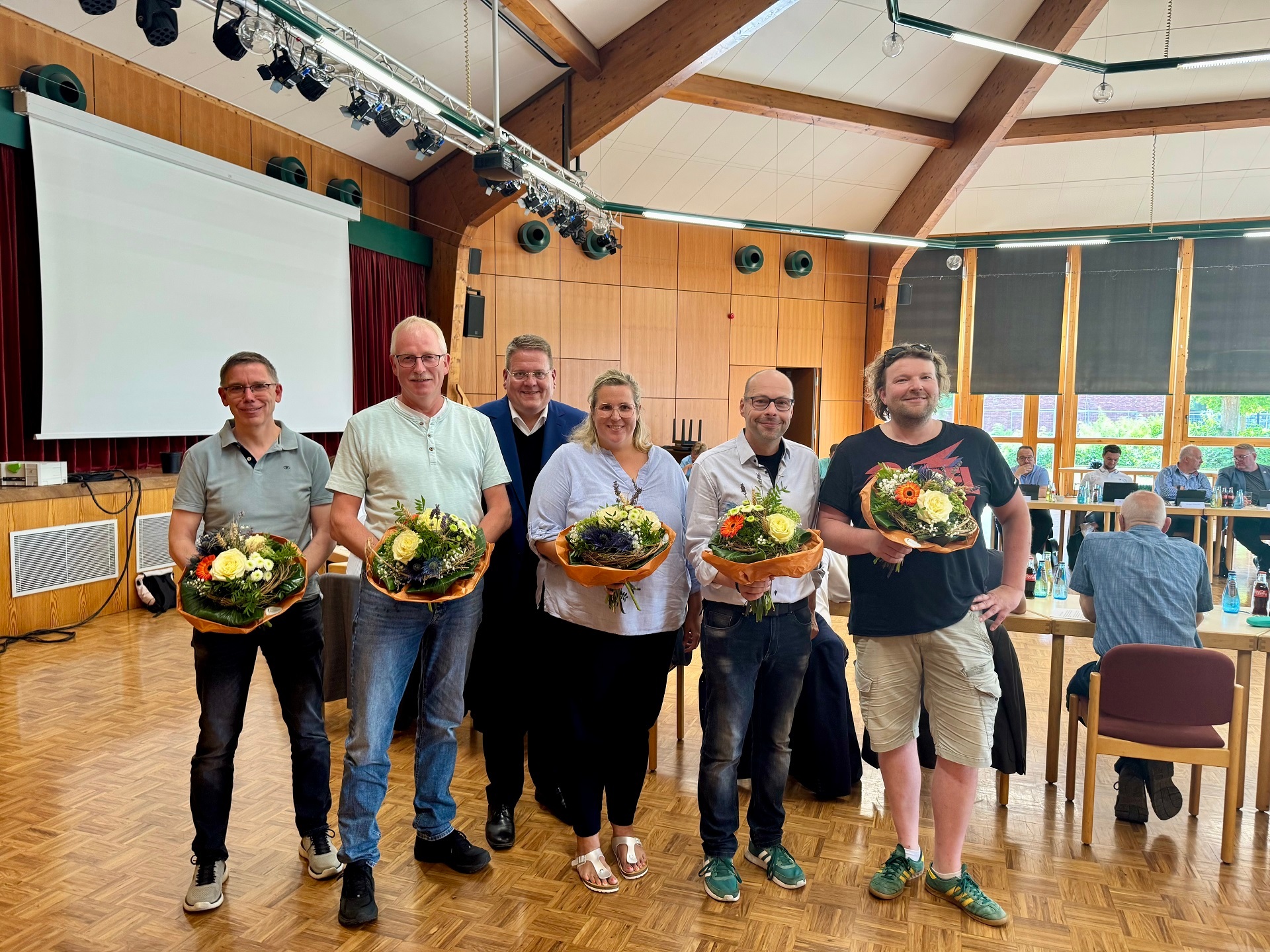 Sechs Personen stehen in einem Saal und halten Blumensträuße, offenbar als Zeichen der Ehrung oder Begrüßung. Im Hintergrund sitzen weitere Menschen an Tischen in einem Versammlungsraum mit Holzdecke und großer Leinwand.