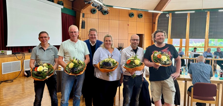 Sechs Personen stehen in einem Saal und halten Blumensträuße, offenbar als Zeichen der Ehrung oder Begrüßung. Im Hintergrund sitzen weitere Menschen an Tischen in einem Versammlungsraum mit Holzdecke und großer Leinwand.