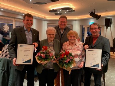 Dulli-Preis 2023 Fünf Personen stehen in einem festlich beleuchteten Raum. Zwei Männer halten gerahmte Urkunden, zwei ältere Frauen halten Blumensträuße. Alle lächeln in die Kamera – offenbar anlässlich einer Ehrung oder Preisverleihung.