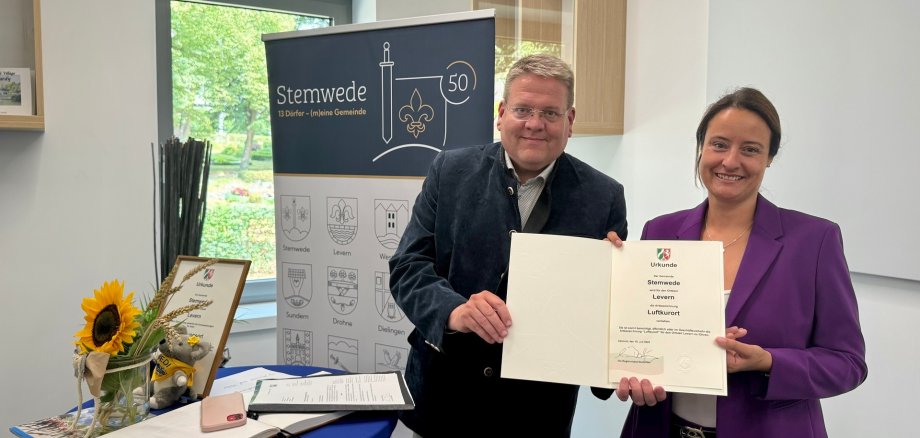 Ein Mann und eine Frau stehen lächelnd in einem Raum und halten gemeinsam eine Urkunde in die Kamera. Neben ihnen steht ein Tisch mit einer Sonnenblume, einem Gästebuch und einem kleinen Stofftier. Im Hintergrund ist ein Banner mit der Aufschrift „Stemwede – 13 Dörfer – (m)eine Gemeinde“ zu sehen.