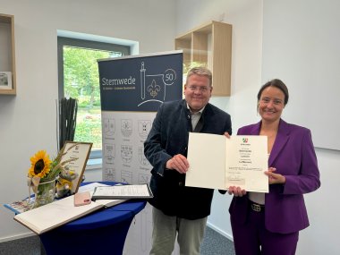 Urkundenübergabe Ein Mann und eine Frau stehen lächelnd in einem Raum und halten gemeinsam eine Urkunde in die Kamera. Neben ihnen steht ein Tisch mit einer Sonnenblume, einem Gästebuch und einem kleinen Stofftier. Im Hintergrund ist ein Banner mit der Aufschrift „Stemwede – 13 Dörfer – (m)eine Gemeinde“ zu sehen.