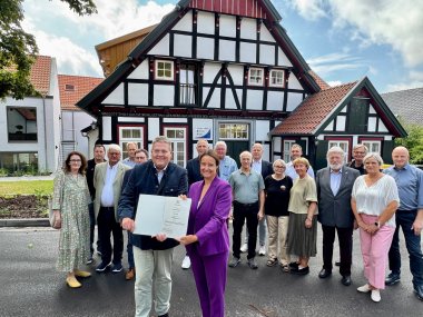 Urkundenübergabe Eine Gruppe von etwa 20 Personen steht vor einem Fachwerkhaus und lächelt in die Kamera. Zwei Personen in der Mitte halten eine Urkunde. Die Szene spielt sich bei sonnigem Wetter im Freien ab.