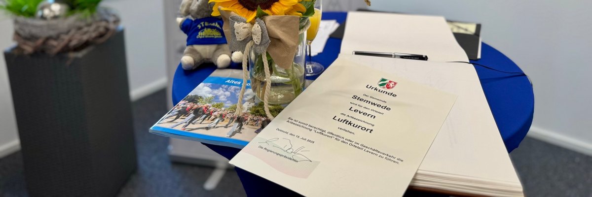 Ein runder Tisch mit einer Urkunde, einer Sonnenblume in einer Vase, einem Stofftier, einem Prospekt und einem offenen Gästebuch. Im Hintergrund ist ein Schild mit der Aufschrift „Stemwede“ und ein Fenster mit Blick ins Grüne zu sehen.