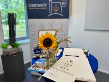 Ein runder Tisch mit einer Urkunde, einer Sonnenblume in einer Vase, einem Stofftier, einem Prospekt und einem offenen Gästebuch. Im Hintergrund ist ein Schild mit der Aufschrift „Stemwede“ und ein Fenster mit Blick ins Grüne zu sehen.