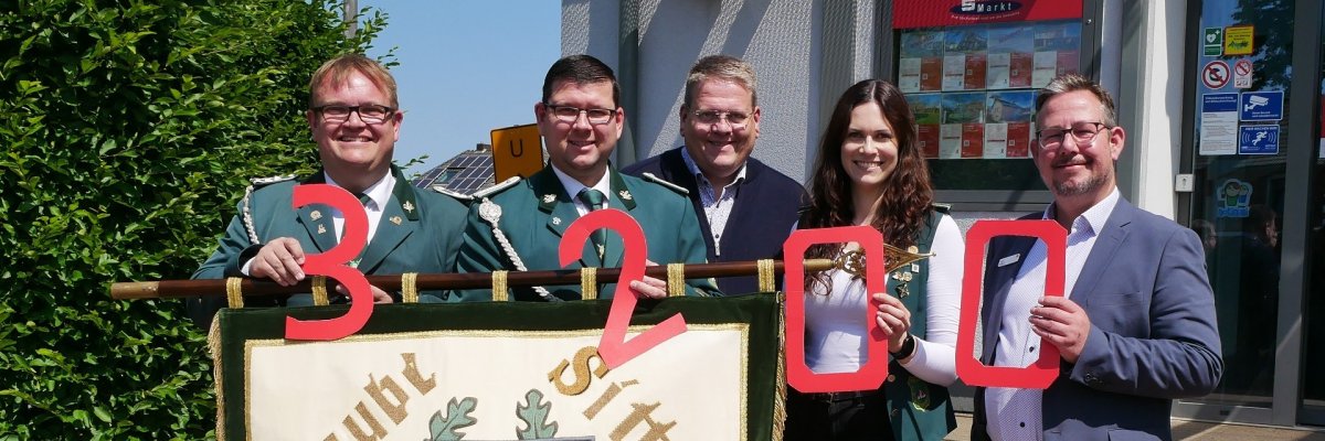 Fünf Personen stehen vor einem Gebäude und halten große rote Zahlen hoch, die zusammen „3200“ ergeben. Davor ist eine Vereinsfahne mit der Aufschrift „Glaube Sitte Heimat“ zu sehen. Drei Personen tragen grüne Uniformen, die anderen drei sind zivil gekleidet.