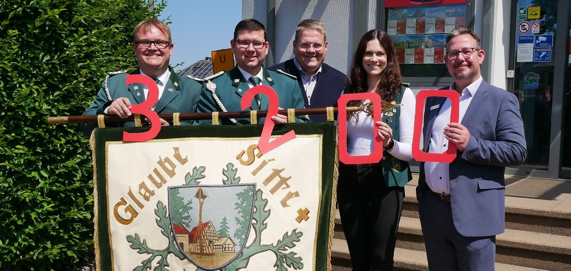 Fünf Personen stehen vor einem Gebäude und halten große rote Zahlen hoch, die zusammen „3200“ ergeben. Davor ist eine Vereinsfahne mit der Aufschrift „Glaube Sitte Heimat“ zu sehen. Drei Personen tragen grüne Uniformen, die anderen drei sind zivil gekleidet.