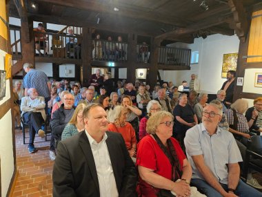 Besuch aus Lardy 2025 Eine Diele eines Fachwerkhauses, in der zahlreiche Menschen sitzen und in Richtung einer Bühne schauen.