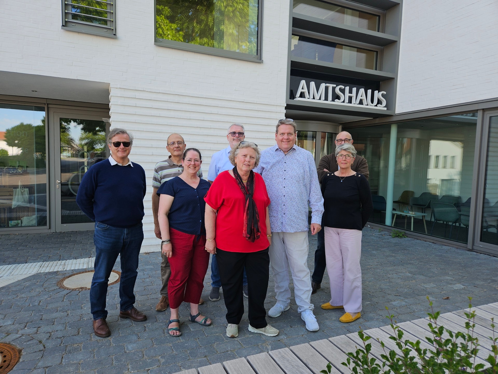 Sieben Menschen posieren vor einem weißen Gebäude, an dessen Fassade die Aufschrift Amtshaus zu lesen ist, für ein Gruppenfoto.